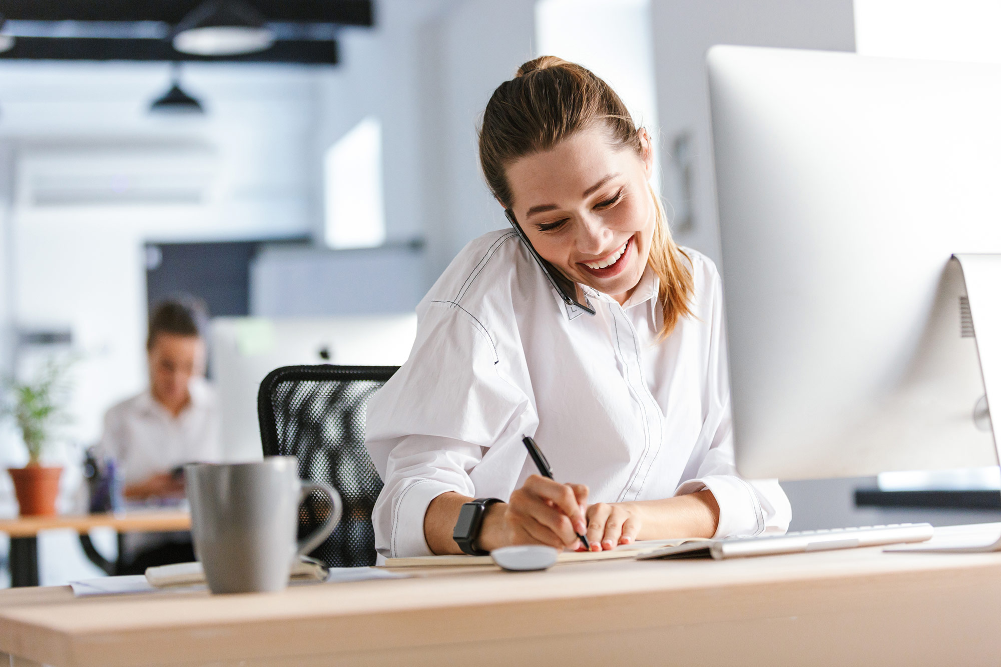 Eine lächelnde Frau telefoniert im Büro und macht gleichzeitig Notizen an ihrem Schreibtisch.