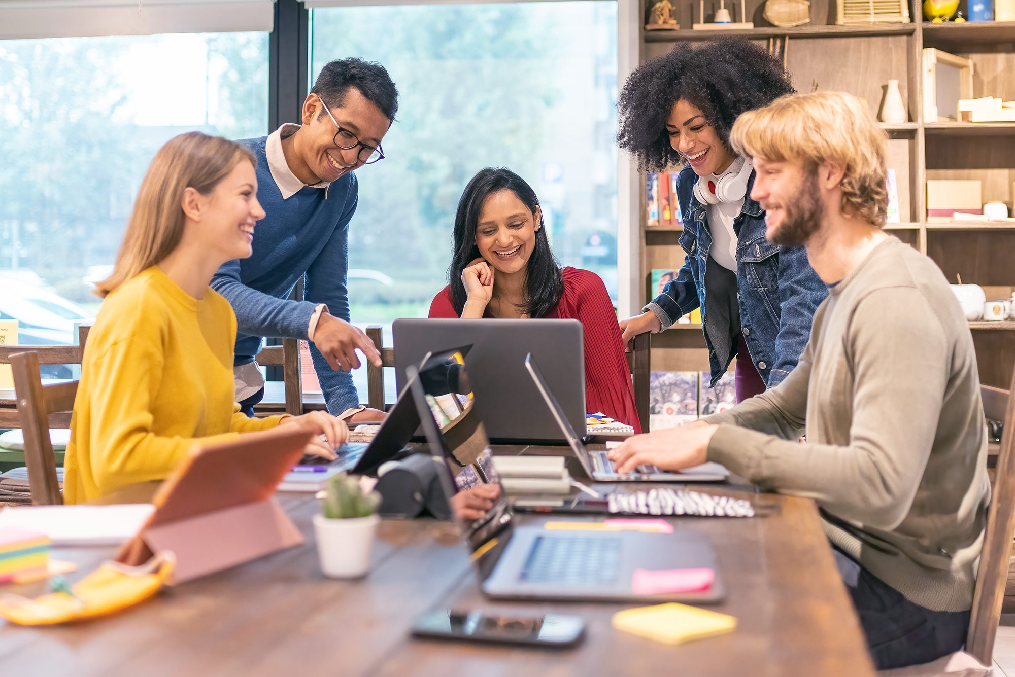 Eine Gruppe von fünf Menschen arbeitet lächelnd gemeinsam an Laptops in einem modernen Büro.