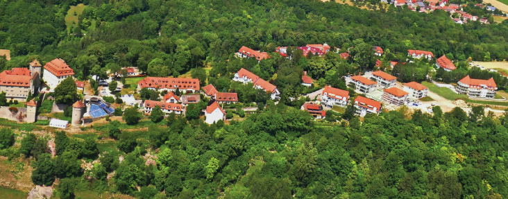 Luftaufnahme eines malerischen Dorfes mit roten Dächern, umgeben von üppigem Grün und einem kleinen Schloss.
